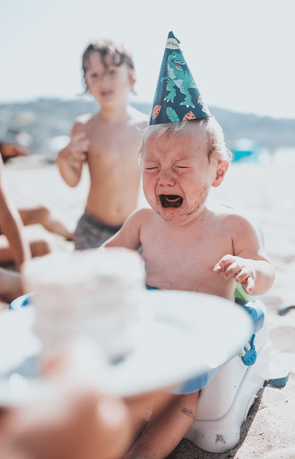 Toddler in a birthday hat cries as a small cake is brought to him on the beach